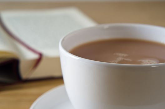 Close up (macro) of a cup of tea with saucer on a wooden table, with an open bookmarked novel in soft focus background.  