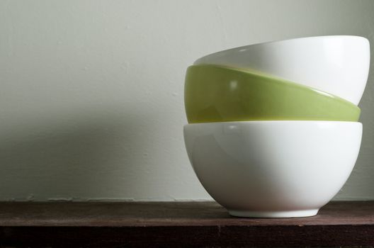 A stack of three china bowls on an old dark wood shelf in a larder with copy space to left on off-white wall. Landscape orientation.