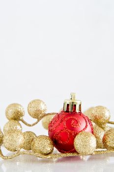 A red Christmas bauble with glittery spiral pattern and gold glitter ball decorations on an off-white reflective surface with copy space above.