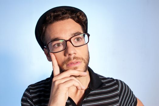 Young man modern nerd thinking with hat and glasses wide angle close up portrait on blue background
