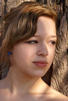 A young attractive woman looks off in the distance while leaning on a tree.