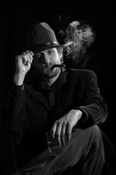 A  man in a  hat sitting with a glass of alcohol and cigar