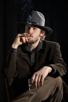A handsome man in a grey hat sitting with a glass of alcohol and cigar