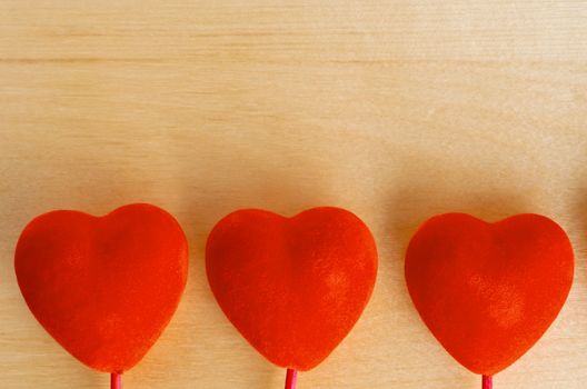 Three velvet-textured red hearts in a row at bottom of frame, with light grained wood background providing copy space above.