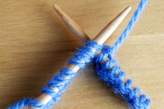 Close up (macro) of pale blue yarn stitches on a pair of wooden knitting needles, laid across eachother on a wooden table.