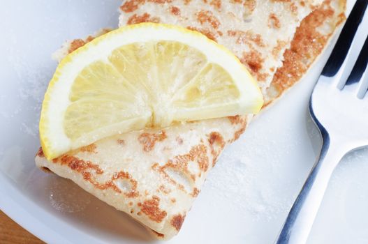 Close up (macro) of a rolled pancake, topped with a lemon slice and sprinkled with caster sugar, on a white plate with a fork.