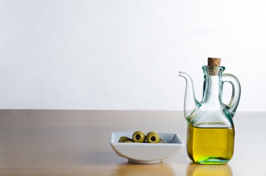 A corked glass jug of olive oil, next to a bowl of olives on a wooden surface.  Off-white background and table provide copy space to left and above.