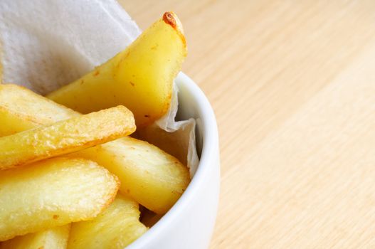 Close up (macro) of a bowl of chips (french fries) nestling on absorbent paper in a white bowl.  Wooden table surface provides copy space to the right.