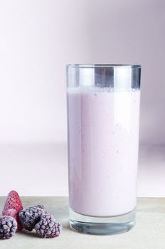A tall glass of pink smoothie on a wooden chopping board with frozen summer fruits, against a pink background.  This is a vegan and dairy-free smoothie made with soya milk.