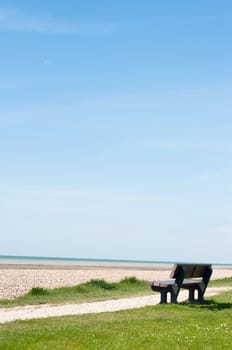 A solitary, empty bench facing the sea on a path between grass and a shingle beach.  A softly clouded blue sky provides two thirds of copy space.