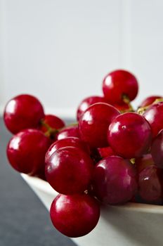 Close up of a bunch of shiny, wet red grapes, tumbling over the edge of a bowl on grey granite kitchen surface with white kitchen tiles in soft focus background.