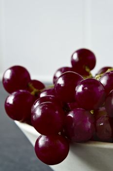 Close up of shiny, wet purple grapes, tumbling over the edge of a bowl on grey granite kitchen surface with white kitchen tiles in soft focus background.  Not their original colour, re-tinted in post-production.