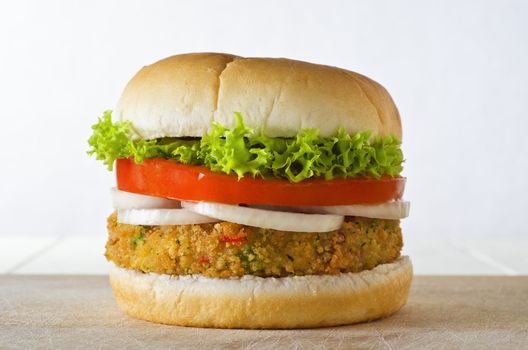 A cheese-free vegetarian burger made from vegetables and breadcrumbs, stacked with onion rings, slice of tomato and curly lettuce, in a bap on a wooden board.