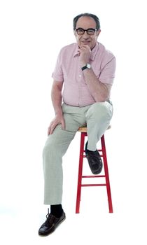 Confident senior man looking at you as he rests on stool, studio shot