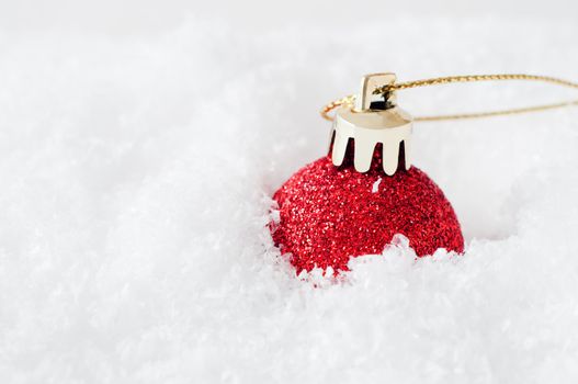 A sparkly red glitter Christmas bauble, with gold clasp and gilt string, sunk into fake white snow to the right of frame.  Snow provides copy space to left.