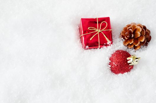 A christmas gift box, bauble and fir cone, embedded in fake white snow to the right of frame, with copy space on the left.