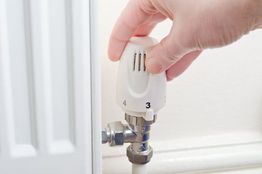 Close up of a hand turning a radiator knob either up or down.  Steel fittings and part of radiator visible, with cream painted wall and skirting board in the background.