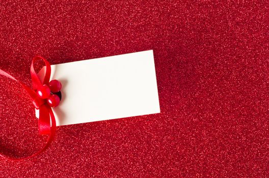 A blank Christmas gift tag, decorated with red ribbon and artifical holly berries, on a sparkly red glitter background.  Copy space on tag.