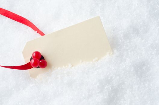 A blank Christmas gift tag, attached to a red ribbon and decorated with plastic red holly berries, nestling in artificial snow.  Copy space on cream coloured tag.