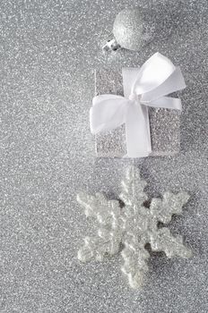 Overhead shot of three silver glittering Christmas ornaments on a silver glitter background. Includes a bauble, a Christmas gift with white ribbon bow, and a star shaped snowflake.  Copy space to left.