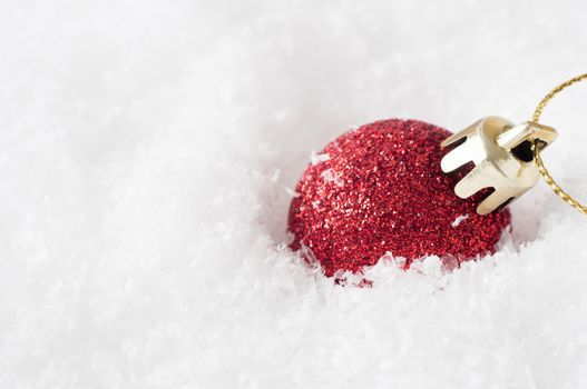 A solitary shiny red Christmas bauble, coated in glitter with gold clasp and string, sunk into artificial white snow and leaning to the right.  Copy space to left.