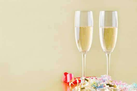 Two fluted champagne glasses with bubbles rising on a gold background with popper streamers and party horn blowers on the surface below.  Copy space to the left.