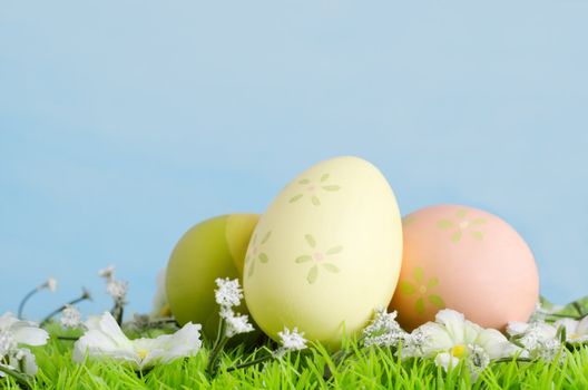 Eggs decoratively painted on artificial grass with fabric flowers photographed against a painted blue background to simulate Spring sky.