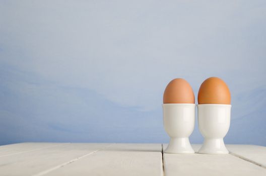 Two fresh brown eggs in egg cups on an old, cream painted wood plank farmhouse table, with painted blue sky effect background.