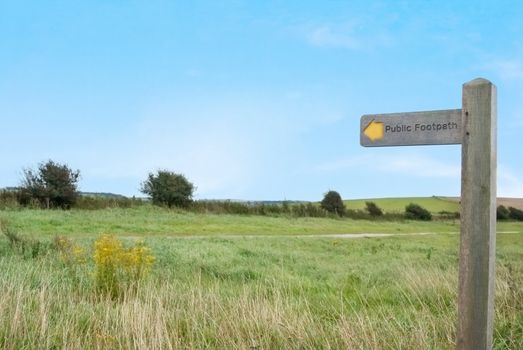 A countryside path under a bright blue sky, surrounded by grass, trees and shrubs on a hilltop.  An arrowed wooden signpost displays 'public footpath' in the foreground, and points towards the path.