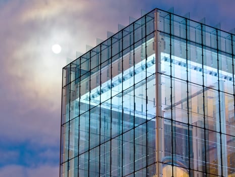 A moon looms over a modernist glass building during the twilight hour.