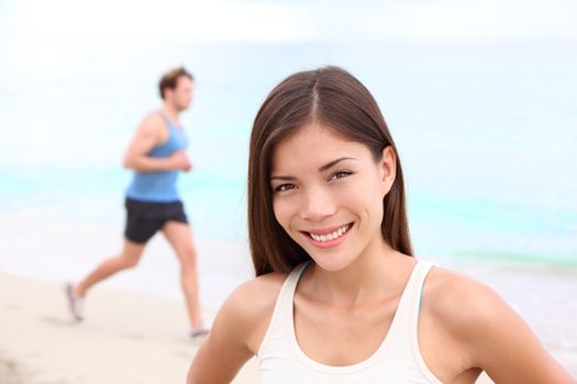 Runner workout woman portrait on beach with man running in background. Happy smiling mixed race Asian / Caucasian female fitness sport model during outdoor workout.