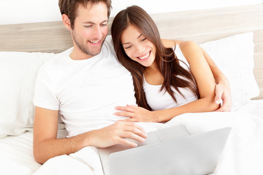 Bed couple with laptop watching movie on computer in bed before sleeping. Happy smiling young interracial couple, Asian woman, Caucasian man in bed at home.
