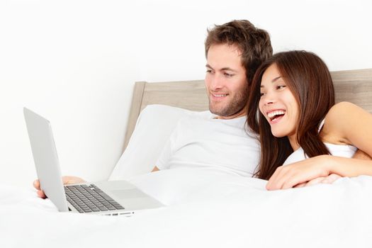 Happy couple with laptop in bed watching fun movie on computer in bed before sleeping. Smiling young interracial couple, Asian woman, Caucasian man in bed at home.