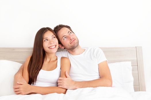 Happy couple in bed thinking and dreaming together looking up at copy space.  Smiling young interracial couple, Asian woman, Caucasian man in bed at home.