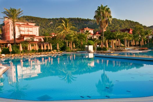 Sweeming pool with blue water near buildings, surrounded by palms, loungers and umbrellas