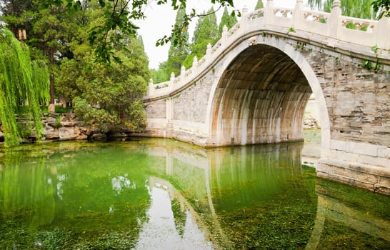 Old style stone Chinese arch bridge in a green garden pond in Beijing, China