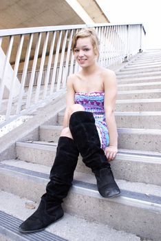 A young adult sits on the concrete stairs in her fashionable dress and boots.