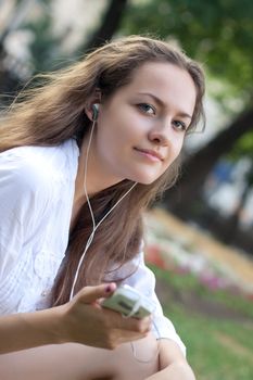 young woman listens to music in ear-phones sits on a lawn in city park