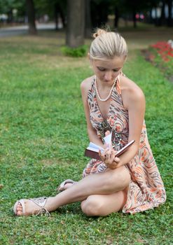 young woman reads the book in hands sits on a green grass in park in a light dress
