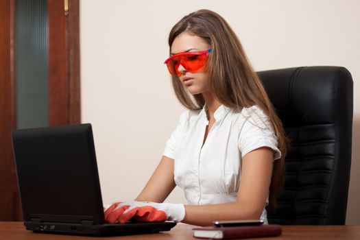 young woman sits at a table at office in orange points and gloves looks in the laptop. Business style