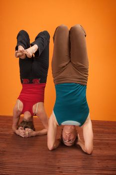 Two women perfrom Vrisikasana yoga posture over orange background