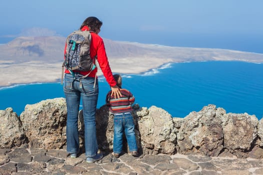 Back view young mother with backpack and son standing on cliff's edge and looking to a island.