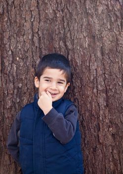 Little boy looking to the left in front of a tree with bark texture in the background biting on his finger with a shy expression.