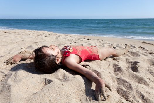 Little girl hanging out on the beach covered in sand. Sleeping girl in red bathing suit on the beach with ocean and sky in the background