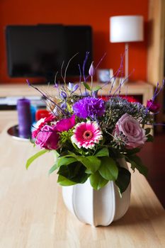 Flower bouquet in vase on table in modern interior with television, lamp and plate with colorful candles in background