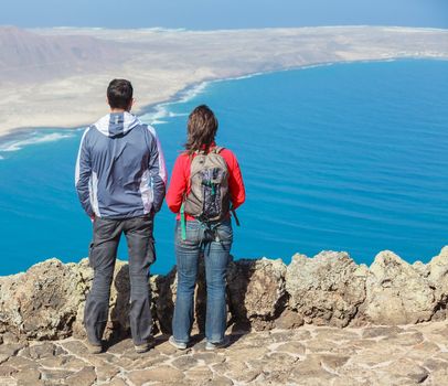 Back view sport couple with backpack standing on cliff's edge and looking to a island.