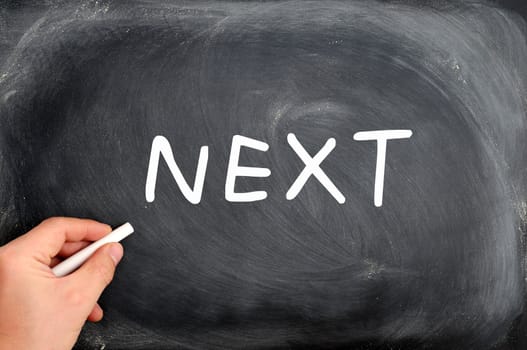NEXT written on a Blackboard background with a hand holding chalk