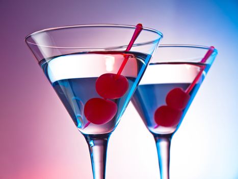 Two glasses of martini with red cherries on a mixed color background