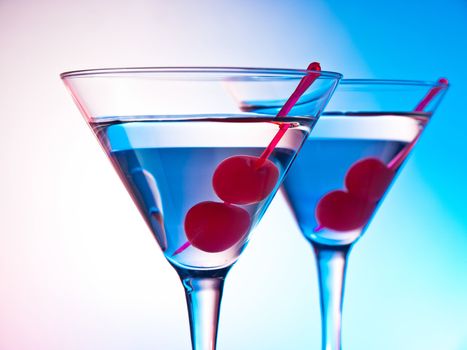 Two glasses of martini with red cherries on a mixed color background