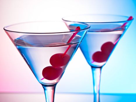 Two glasses of martini with red cherries on a mixed color background